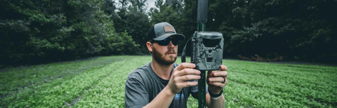 Man attaching Spartan GoLive trail camera to a post in a field surrounded by trees.
