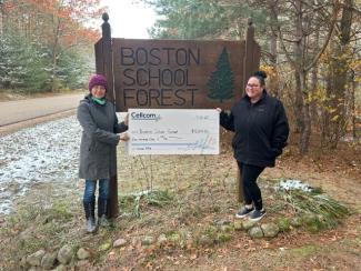 Two people holding a donation check in front of the Boston School Forest sign.