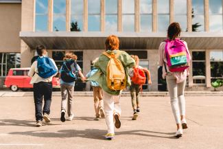Group of six kids with backpacks walking into a school on a sunny day.